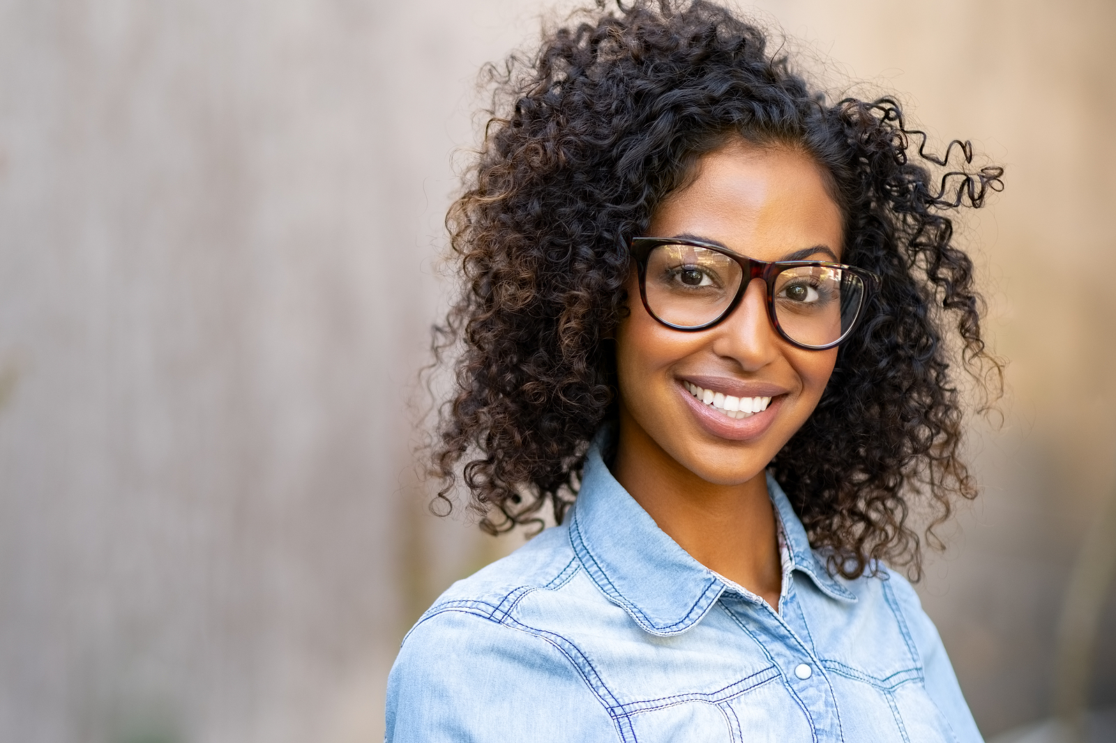 Happy smiling young woman with eyeglasses and denim shirt looking at camera. Black girl feeling successful standing against wall with copy space. African student smiling and wearing spectacles.